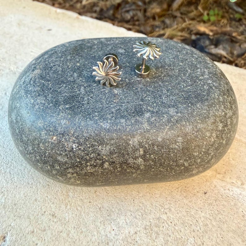 May include: Two silver-toned stud earrings featuring a sunburst design with radiating rays. The earrings have a metallic finish and a post-back mechanism, displayed on a smooth, dark gray, oval-shaped stone surface.