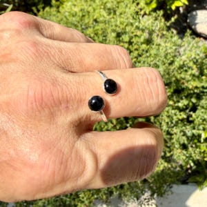 May include: A silver open-style ring featuring two polished black spherical stones at each end. The band is crafted from a smooth, reflective silver-toned metal, and the two black beads are set symmetrically to create a bypass design.