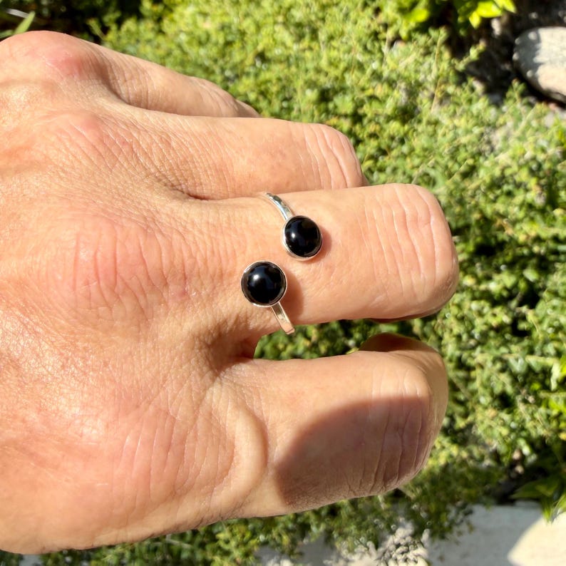 May include: A silver open-style ring featuring two polished black spherical stones at each end. The band is crafted from a smooth, reflective silver-toned metal, and the two black beads are set symmetrically to create a bypass design.