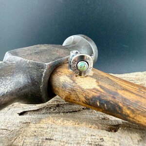 May include: A silver ring featuring a turquoise stone set within a bullet casing. The ring is engraved with the numbers "40" and the letters "S&W" and "WLLN". The ring is displayed on a wooden surface.