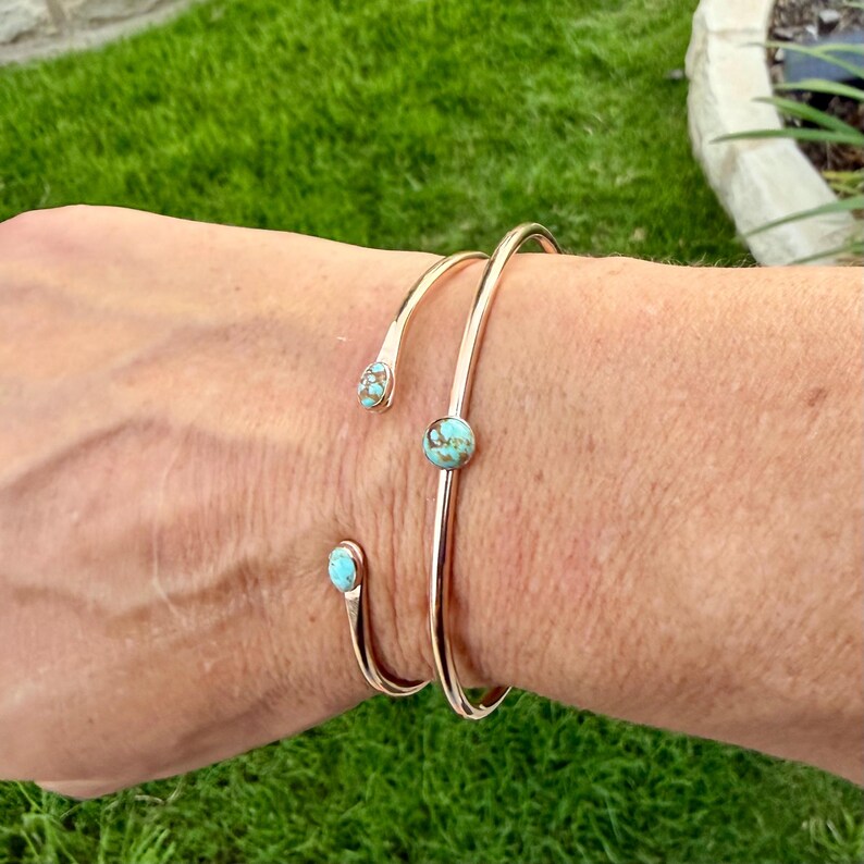 May include: Two gold-colored open cuff bracelets with turquoise stones. One bracelet has a single turquoise stone in the center, while the other has two turquoise stones at the ends. The bracelets are worn on a person's arm.