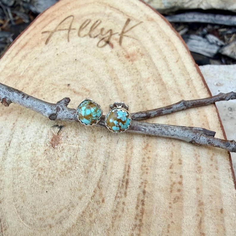 May include: A pair of turquoise and brown stone stud earrings with a silver-colored scalloped edge. The earrings are displayed on a small branch, resting on a wooden surface. The name "Ally K" is etched into the wood.
