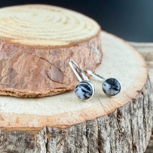 May include: A pair of silver drop earrings with round, black and white patterned stones. The earrings are displayed on a rustic wooden surface, showcasing their natural stone design and elegant style.