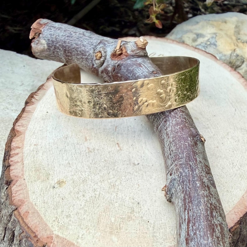 Puede incluir: Una pulsera tipo brazalete ancha de metal dorado con una superficie texturizada y martillada que presenta un patr&oacute;n org&aacute;nico irregular en toda la banda exterior. La pulsera es abierta y mantiene un ancho constante en su dise&ntilde;o.