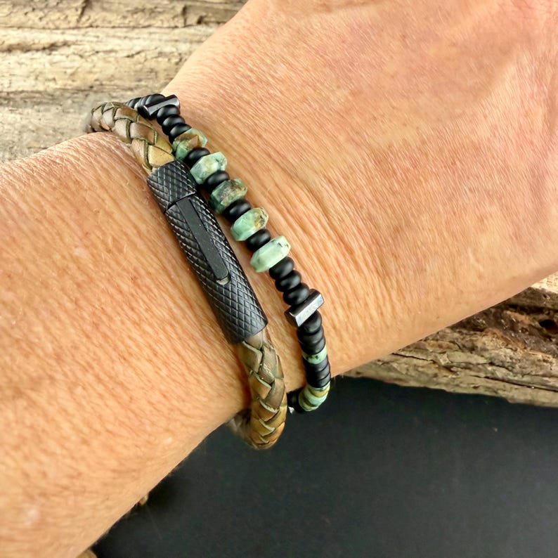 May include: Two bracelets are shown. One is a braided leather bracelet with a black clasp. The other bracelet is made of black beads and turquoise-colored stones. The bracelets are worn on a wrist.