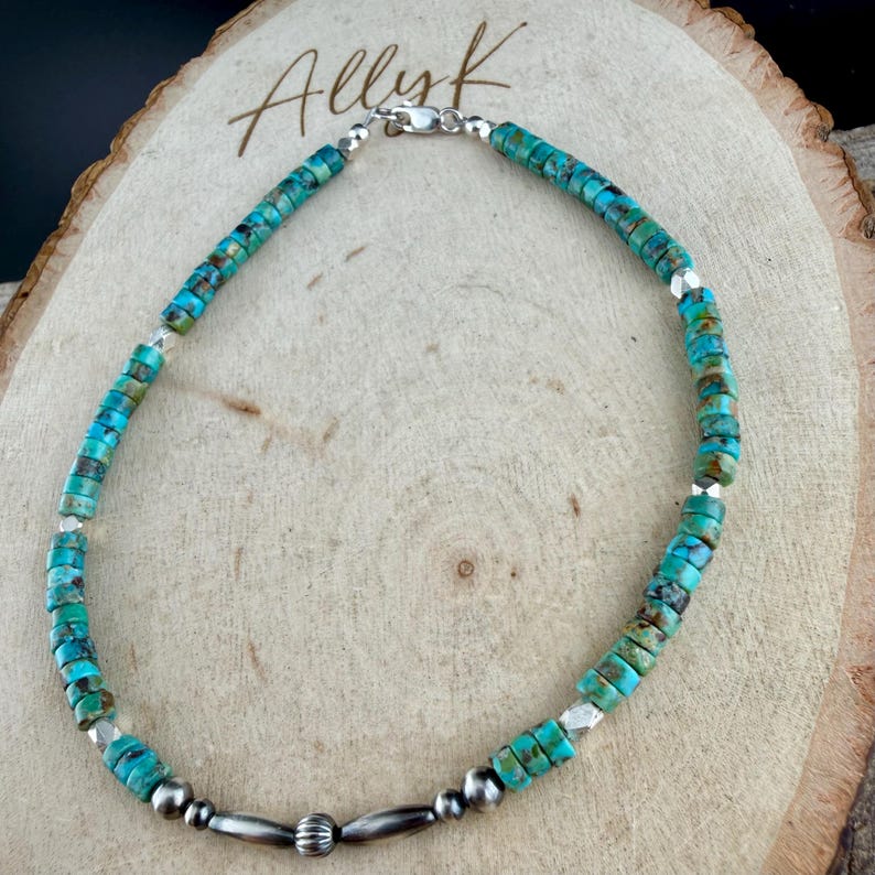 Pu&ograve; includere: Collana di perline turchesi con dettagli argentati. La collana presenta piccole perline rotonde turchesi in tonalit&agrave; blu e verde, infilate con perline di metallo argentato e una chiusura. La collana &egrave; esposta su una superficie di legno.