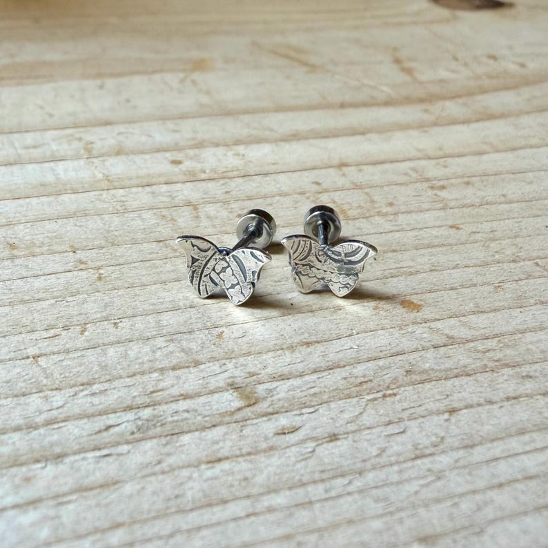 K&ouml;nnte beinhalten: Ein Paar silberfarbene Ohrstecker in Schmetterlingsform. Jeder Ohrstecker weist eine detaillierte, texturierte Oberfl&auml;che mit komplexen Linienmustern auf den Fl&uuml;geln sowie einen kleinen Stiftverschluss zur Befestigung am Ohrl&auml;ppchen auf.