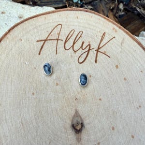 May include: Oval-shaped stud earrings with a silver-colored border, featuring a dark blue and gray marbled design. The earrings are displayed on a wooden surface with the handwritten text "Allyk".