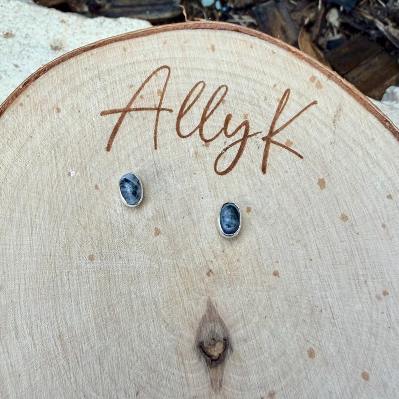 May include: Oval-shaped stud earrings with a silver-colored border, featuring a dark blue and gray marbled design. The earrings are displayed on a wooden surface with the handwritten text "Allyk".