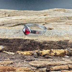 May include: A silver ring with a square, deep red gemstone. The band features etched detailing. The ring is displayed on a textured, brown wooden surface, showcasing the jewelry's craftsmanship and design.