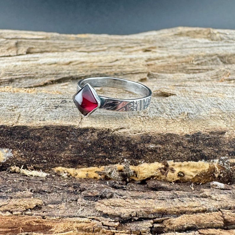 May include: A silver ring with a square, deep red gemstone. The band features etched detailing. The ring is displayed on a textured, brown wooden surface, showcasing the jewelry's craftsmanship and design.
