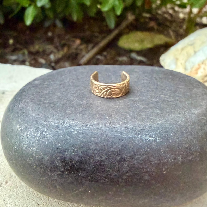 May include: A gold-colored ear cuff with an ornate, engraved design. The jewelry piece is open at the top and bottom, and sits on a dark gray stone. The background is blurred with green foliage.