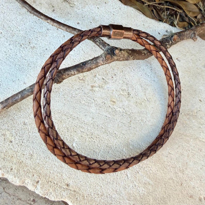 Puede incluir: Un collar de cord&oacute;n de cuero trenzado marr&oacute;n con un cierre magn&eacute;tico cil&iacute;ndrico de metal en tono cobre. El cord&oacute;n de cuero est&aacute; tejido en forma circular, mostrando una variaci&oacute;n de color natural entre marr&oacute;n claro y oscuro en el trenzado.