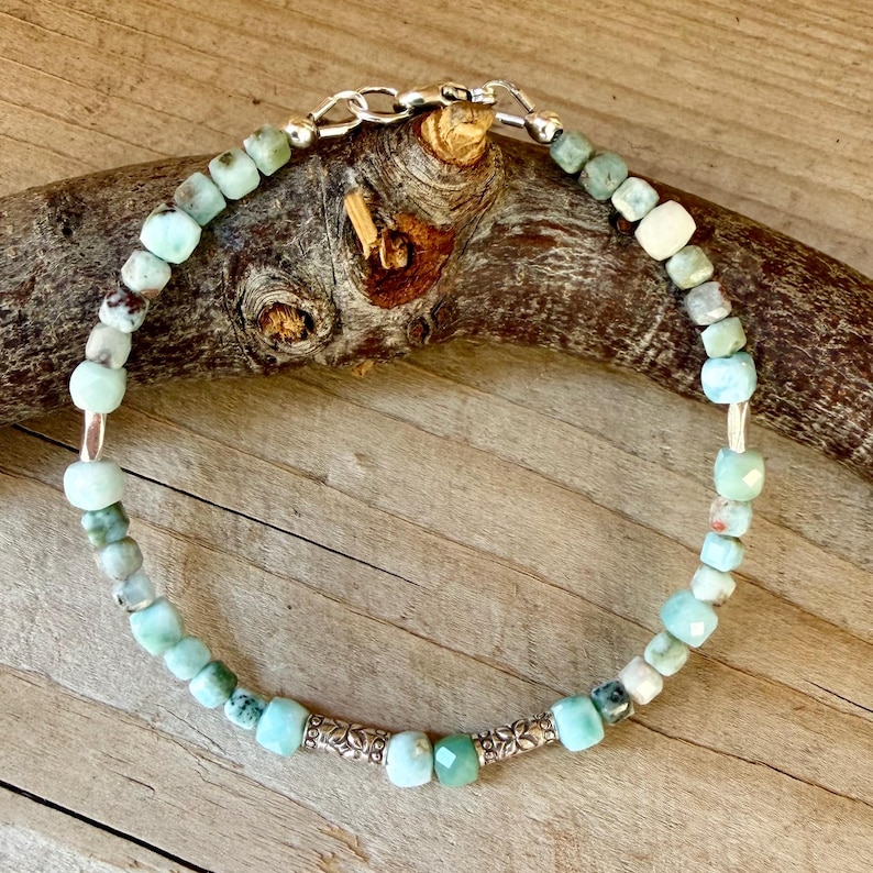 Puede incluir: Una pulsera de cuentas con peque&ntilde;as piedras facetadas de color azul claro y verde, intercaladas con dos espaciadores cil&iacute;ndricos de metal plateado con un patr&oacute;n floral grabado. La pulsera se cierra con un broche de langosta de metal plateado.