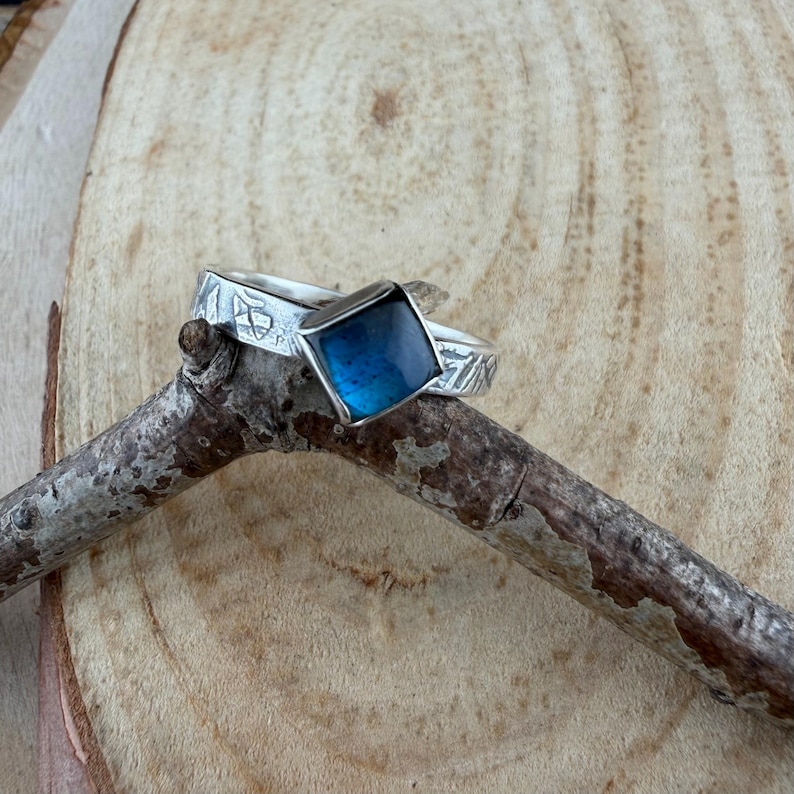 May include: A silver ring with a square, blue gemstone. The band has etched designs. The ring is displayed on a small, brown branch against a wooden background. The gemstone has a dark blue color.