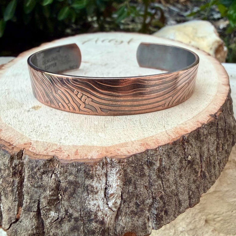 May include: A copper cuff bracelet with a textured, wave-like pattern. The bracelet is a warm copper color with a darker interior and the words "GENUINE COPPER" stamped inside. The bracelet is displayed on a wooden surface.