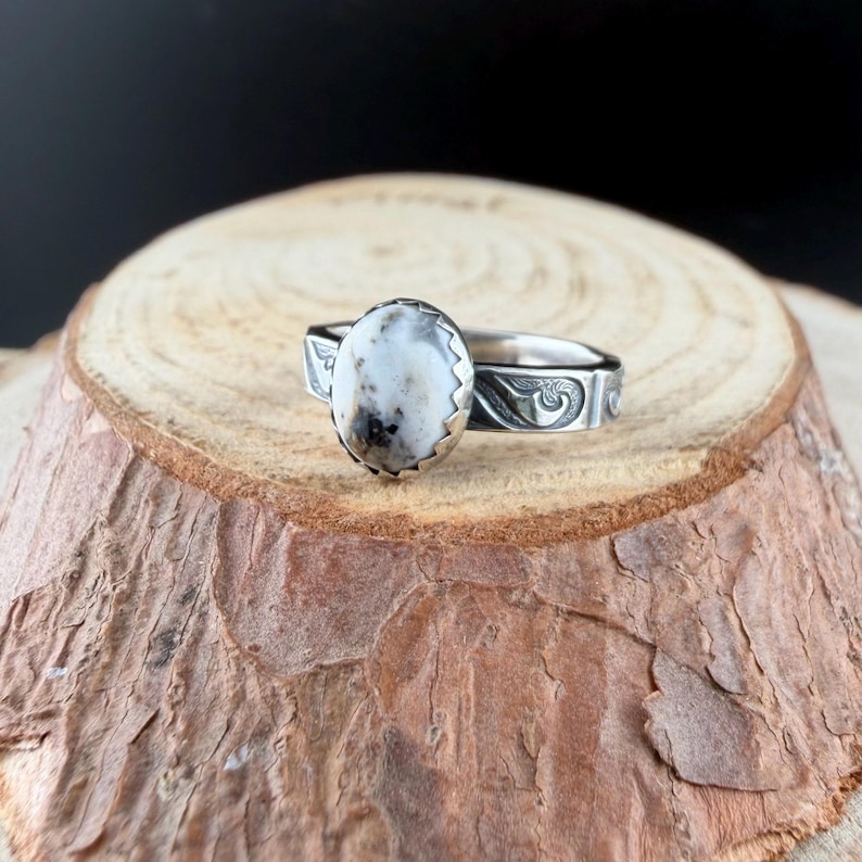 May include: A silver ring with an oval, white and gray stone. The stone is set in a serrated bezel. The band features an engraved, swirling design. The ring is displayed on a wooden surface.