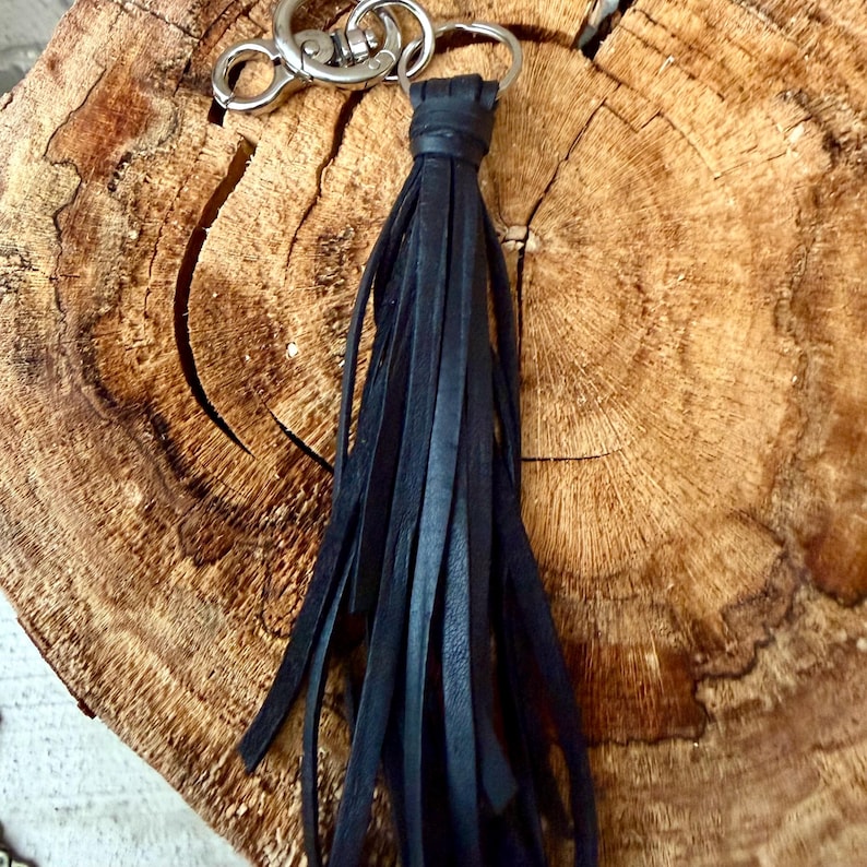 May include: A black leather tassel keychain with a silver-colored clasp and ring. The tassel is made of long, thin strips of black leather, and the keychain is displayed on a textured, brown wooden surface.