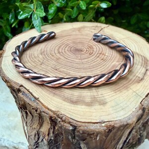 May include: A copper-colored cuff bracelet with a twisted rope design. The bracelet is displayed on a wooden surface with visible growth rings. The metal has a polished finish, reflecting light. The overall aesthetic is rustic and handcrafted.