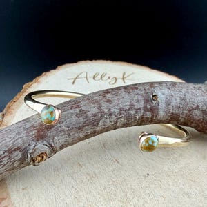 May include: A gold-colored cuff bracelet with turquoise-colored stones at each end. The bracelet is resting on a piece of wood. The word "Allyk" is written on the wood.