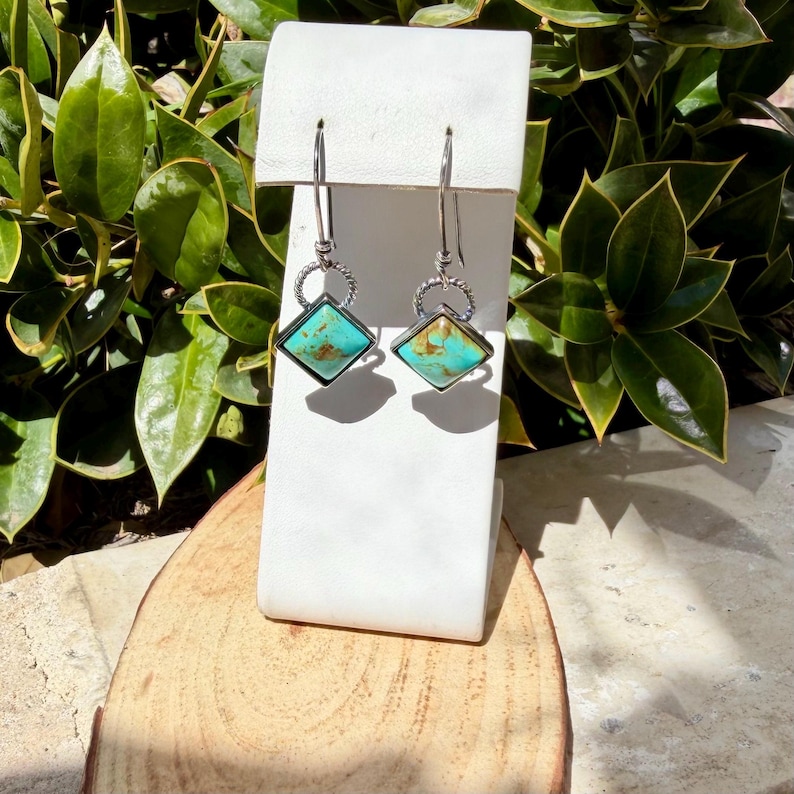 May include: A pair of turquoise and silver earrings. Each earring features a square turquoise stone set in a silver frame, with a decorative silver ring above the stone. The earrings hang from a silver hook.