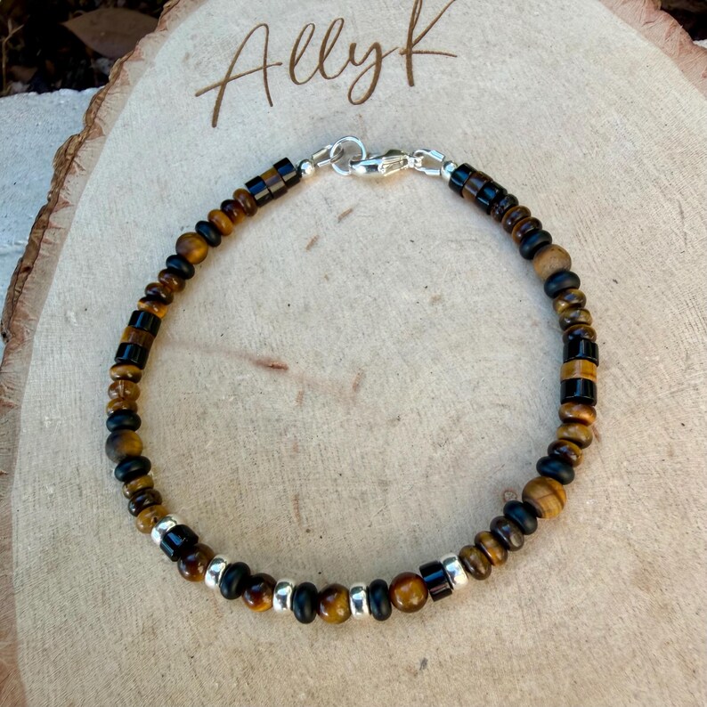 K&ouml;nnte beinhalten: Ein Perlenarmband mit abwechselnden runden Tigerauge-Perlen, zylindrischen schwarzen Perlen und kleinen silberfarbenen Metall-Abstandshaltern. Das Armband ist mit einem silberfarbenen Karabinerverschluss versehen und kreisf&ouml;rmig angeordnet.