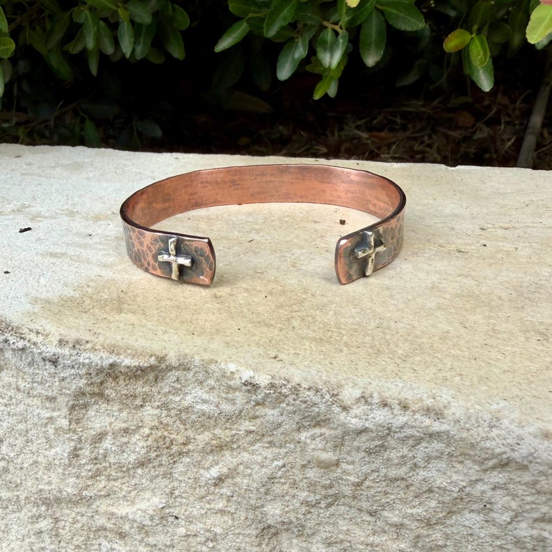 May include: A copper cuff bracelet with a hammered texture and an open design. The bracelet features a small, raised silver-toned cross emblem on each of the two ends. The metal has a warm, reddish-brown finish with darkened, oxidized accents.