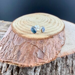 May include: A pair of oval-shaped stud earrings with a silver post. One earring features a dark blue and green stone, while the other has a light blue stone. The earrings are displayed on a wooden surface.