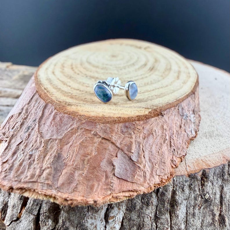 May include: A pair of oval-shaped stud earrings with a silver post. One earring features a dark blue and green stone, while the other has a light blue stone. The earrings are displayed on a wooden surface.