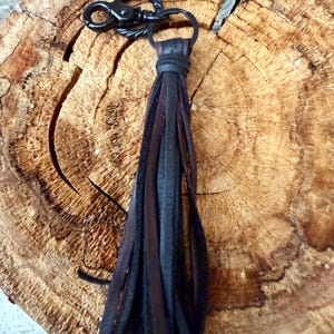 May include: A leather tassel keychain with a black metal clasp and ring. The tassel features strips of black, dark brown, and medium brown leather. The keychain is displayed on a textured, wooden surface.