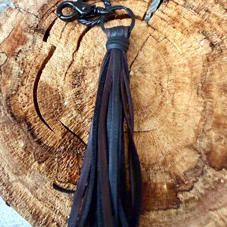 May include: A leather tassel keychain with a black metal clasp and ring. The tassel features strips of black, dark brown, and medium brown leather. The keychain is displayed on a textured, wooden surface.