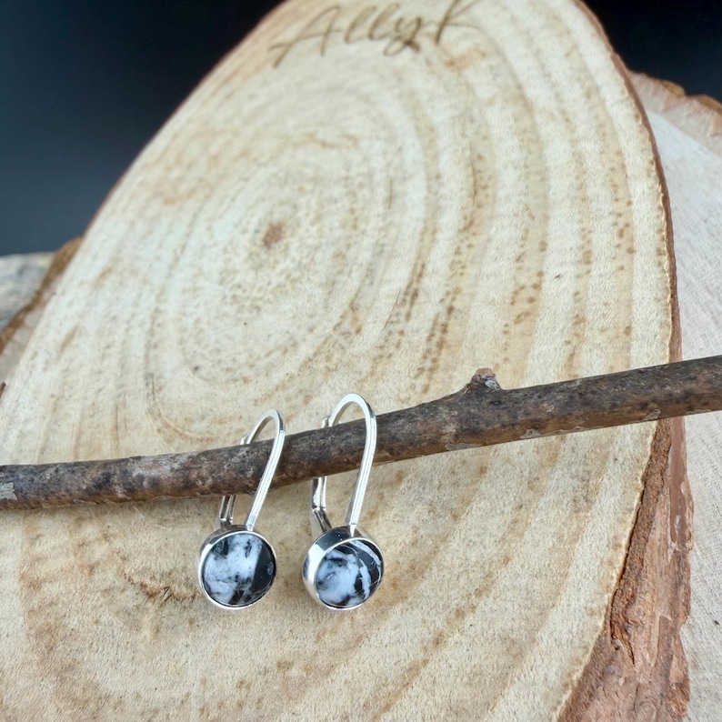 May include: A pair of silver earrings with round, black and white patterned stones. The earrings are displayed on a wooden surface with the handwritten word "Allyk" visible in the background.