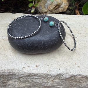May include: A pair of hoop earrings featuring a continuous beaded silver-tone metal texture. Each earring is accented with a single round turquoise stone bead attached to the top of the hoop, showcasing natural blue and brown veining patterns.