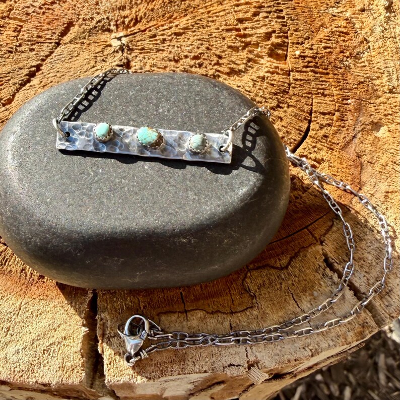 May include: A silver necklace featuring a hammered bar pendant with three turquoise stones. The pendant is connected to a delicate chain with a lobster clasp. The necklace is displayed on a dark gray stone against a wood background.
