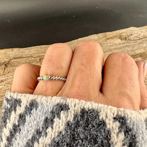 May include: A silver-colored ring with a twisted band and a small, round turquoise and gold-colored stone. The ring is on a finger, and the person is wearing a gray and white patterned sweater.