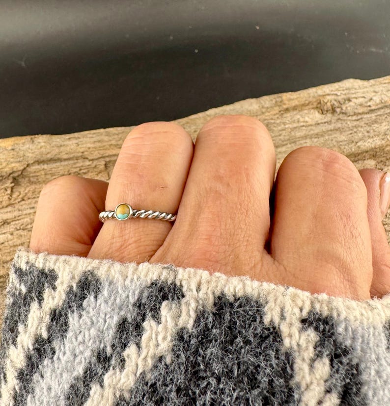 May include: A silver-colored ring with a twisted band and a small, round turquoise and gold-colored stone. The ring is on a finger, and the person is wearing a gray and white patterned sweater.