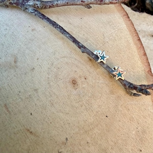 May include: A pair of star-shaped stud earrings with a silver-colored outline and a teal-colored center. The earrings are displayed on a dark brown twig, set against a light brown wooden surface.