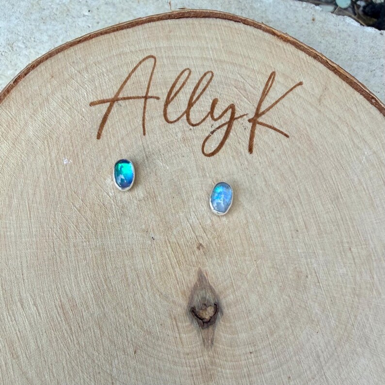 May include: A pair of oval-shaped stud earrings with iridescent blue and green stones set in silver. The earrings are displayed on a wooden surface with the name "Allyk" engraved on it.