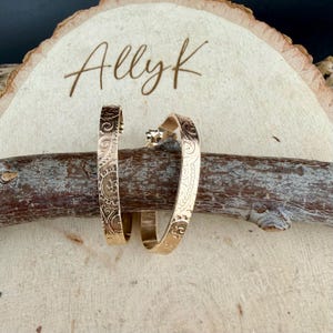 May include: A pair of gold-colored hoop earrings with an engraved floral design. The earrings are displayed on a wooden log, with the name "Ally K" written on a wooden slice in the background.