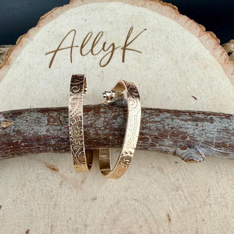 May include: A pair of gold-colored hoop earrings with an engraved floral design. The earrings are displayed on a wooden log, with the name "Ally K" written on a wooden slice in the background.