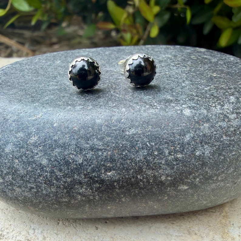 May include: A pair of black onyx stud earrings. Each earring features a round, polished black stone set in a silver-colored, scalloped bezel. The earrings are displayed on a gray, textured stone surface.
