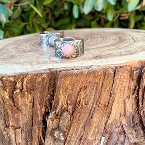 May include: Two silver rings featuring a textured, etched surface and a central round, smooth, pale pink stone. The rings have an open-band design with a scalloped metal setting surrounding the pink cabochon, showcasing a metallic finish.