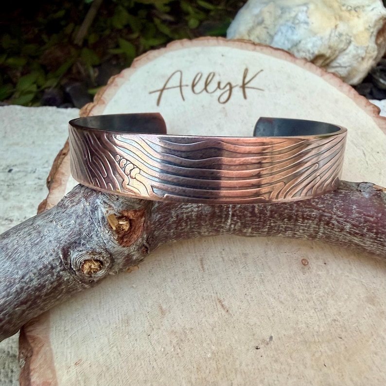 May include: A copper cuff bracelet with a textured, wave-like pattern. The bracelet is open at the bottom and features the name "Ally K" on the wooden surface behind it. The metal has a gradient of colors, from dark to light.
