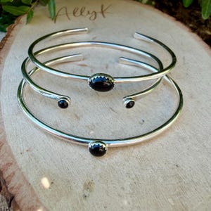 Op de afbeelding: Drie zilverkleurige armbanden met zwarte edelsteenaccenten. E&eacute;n armband heeft een ovale steen, de andere ronde stenen. De armbanden worden gepresenteerd op een houten oppervlak, met de handgeschreven tekst "Allyk" zichtbaar.