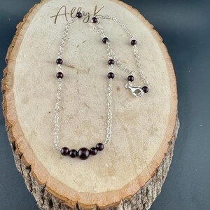 May include: A silver chain necklace with dark red garnet beads. The necklace features a series of small beads along the chain, with a cluster of larger beads at the center. The clasp is visible. The necklace is displayed on a wooden surface.
