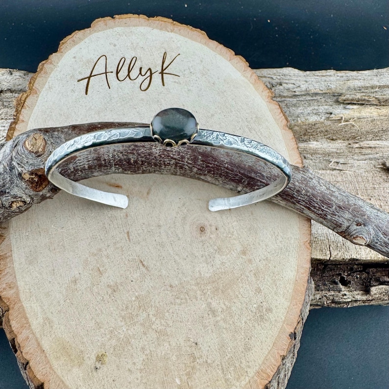 May include: A silver cuff bracelet with an oval, dark gemstone set in a decorative bezel. The bracelet has an open design and is engraved with a floral pattern. The name "Allyk" is written in cursive on the wooden surface.