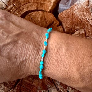 May include: A delicate bracelet featuring small, irregular, bright turquoise-colored stone beads alternating with thin, cylindrical silver-toned metal spacers, creating a simple and linear design worn around the wrist.