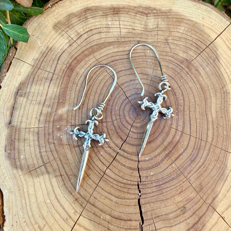 May include: A pair of silver-toned earrings featuring a sword-shaped pendant with an ornate cross-guard design. Each earring hangs from a curved wire hook, showcasing a polished metallic finish with intricate detailing on the cross-shaped hilt.
