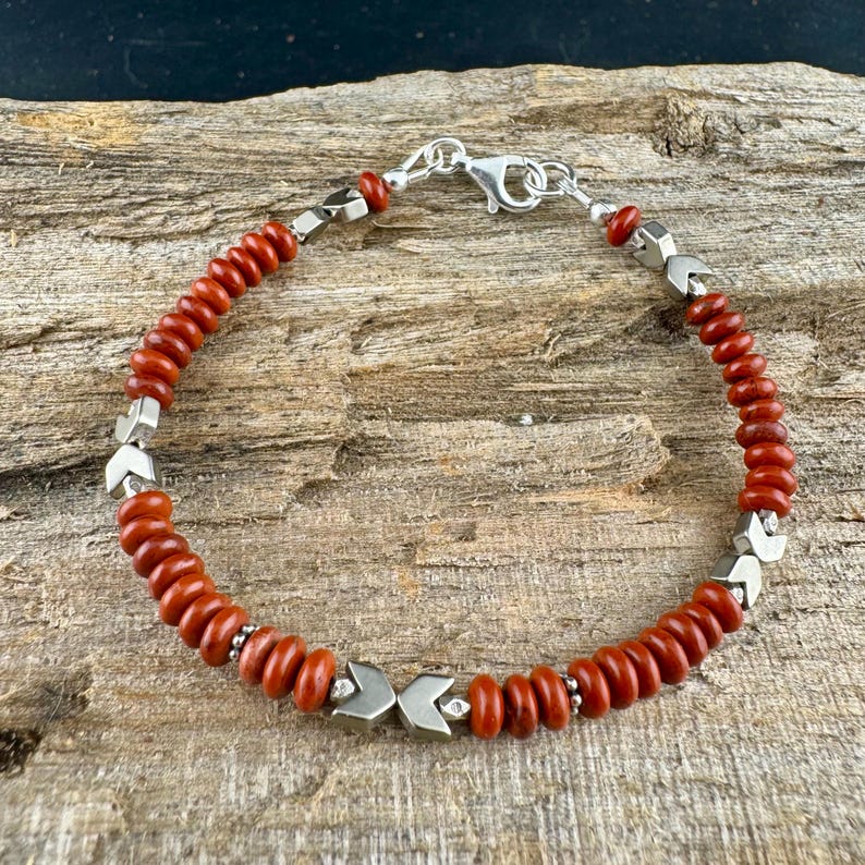 May include: A beaded bracelet featuring small, round, reddish-brown beads interspersed with silver-colored geometric accents. The bracelet has a silver clasp and is displayed on a wooden surface.
