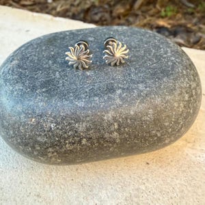 May include: A pair of silver-toned stud earrings featuring a sunburst design with multiple radiating rays. The earrings are metallic with a polished finish and are shaped like a stylized sun with a central point and curved, tapered rays.